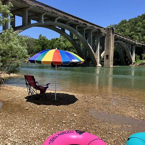 A colorful umbrella and a pink chair sit on a rocky riverbank under a large arched concrete bridge, with bright float tubes nearby—perfect for guests from Cabin Rentals and RV Park in Galena MO to enjoy the scenery on a sunny day.