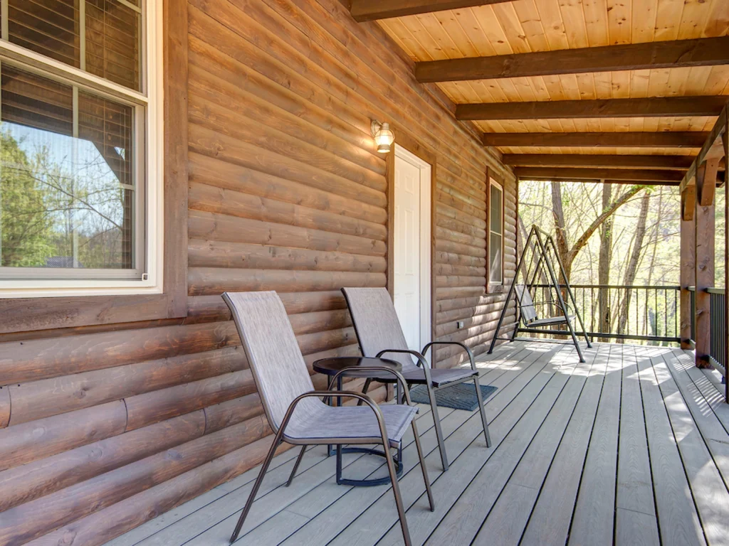 A wooden cabin porch with two lounge chairs, a small table, a swing, and a door; sunlight filters through trees in the background, creating a peaceful, rustic atmosphere perfect for Cabin Rentals and RV Park in Galena MO.