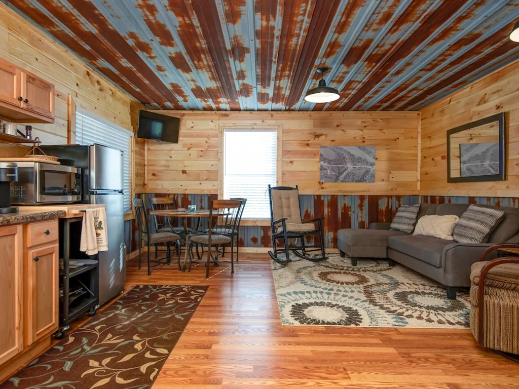 A cozy cabin living area with wood-paneled walls, rustic ceiling, small kitchen, dining table, gray sofa, rocking chairs, patterned rugs, and a large window letting in natural light—perfect for Cabin Rentals and RV Park in Galena MO.