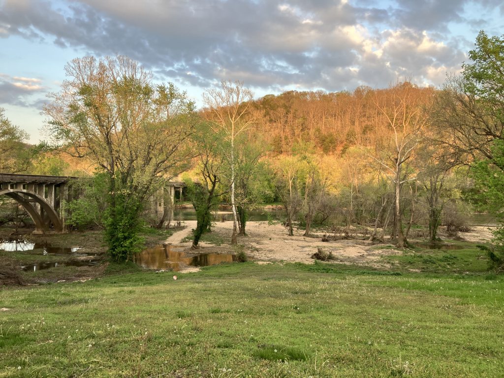 A grassy field leads to a sandy riverbank with scattered trees, an old concrete bridge, and wooded hills in the background—perfect scenery for Cabin Rentals and RV Park in Galena MO under a partly cloudy sky at sunset.