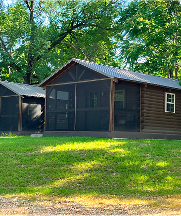 Two small, dark brown, log-style cabins with screened porches sit on a grassy lawn surrounded by tall green trees and dappled sunlight—perfect for cabin rentals near James River MO.