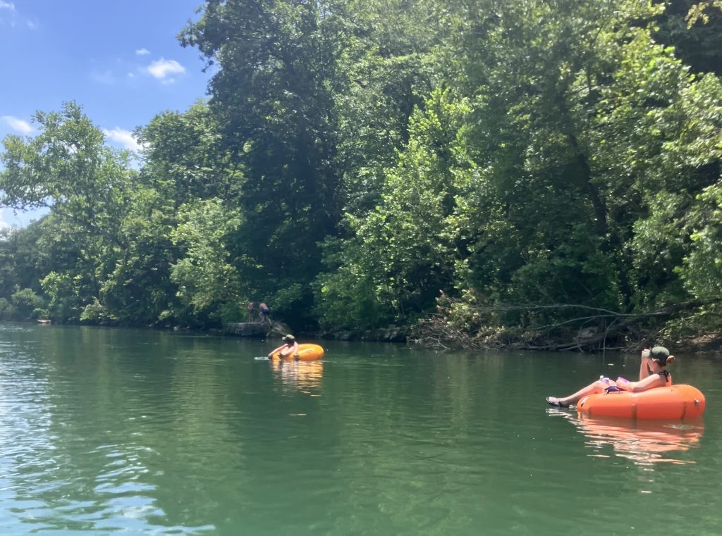 Two people float on orange inner tubes in a calm, green river surrounded by dense, leafy trees under a blue sky near cozy Cabin Rentals and RV Park in Galena MO.
