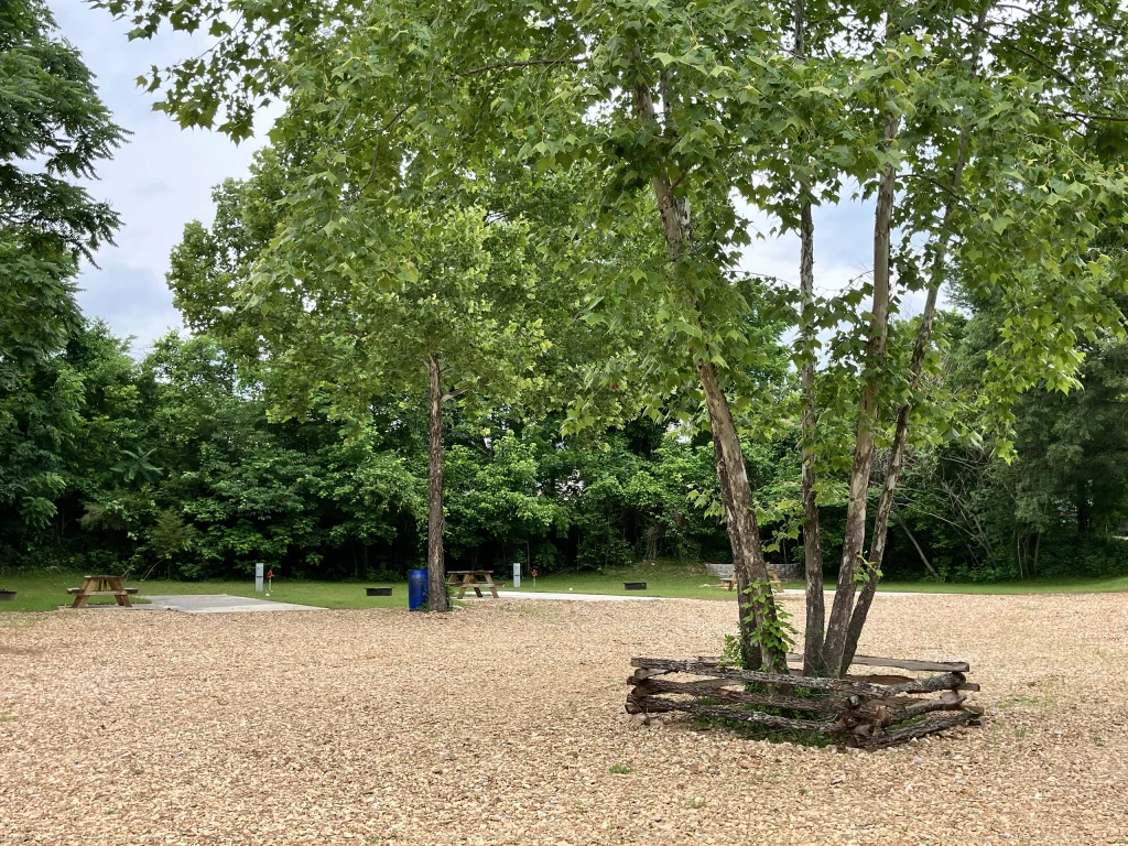 A tree surrounded by a wooden fence stands on a gravel area at our Cabin Rentals and RV Park in Galena MO, with picnic tables and green trees in the background under a partly cloudy sky.