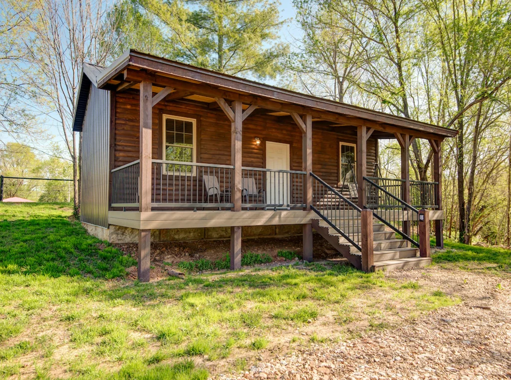 A small wooden cabin with a covered front porch, several chairs, steps leading up to the entrance, and surrounded by green grass and trees on a sunny day—perfect for Cabin Rentals and RV Park in Galena MO.