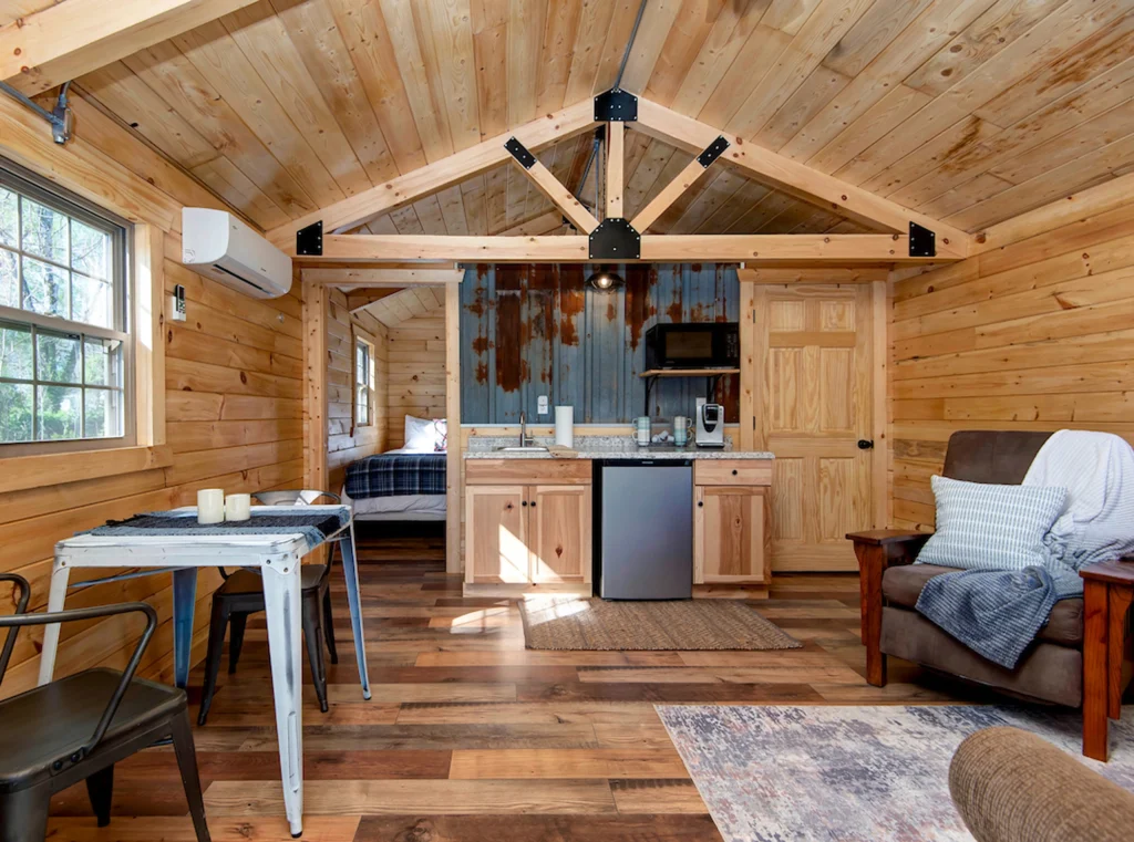 A cozy cabin interior at Cabin Rentals and RV Park in Galena MO features wood-paneled walls, a small kitchenette, a comfy bed, a table with chairs, and an armchair. Natural light pours in through the windows for a warm, welcoming vibe.